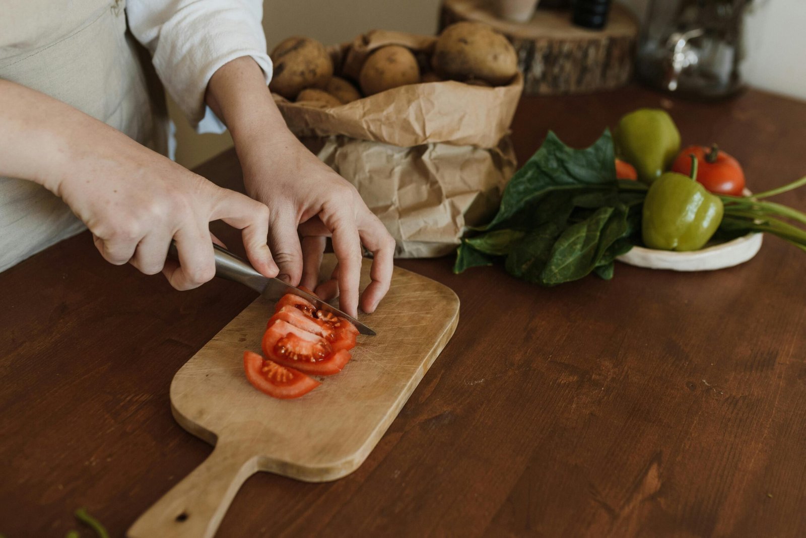 A person is slicing a tomato on a wooden cutting board using a kitchen knife. In the background, there is a paper bag filled with potatoes and a small plate with green bell peppers, spinach, and a tomato.