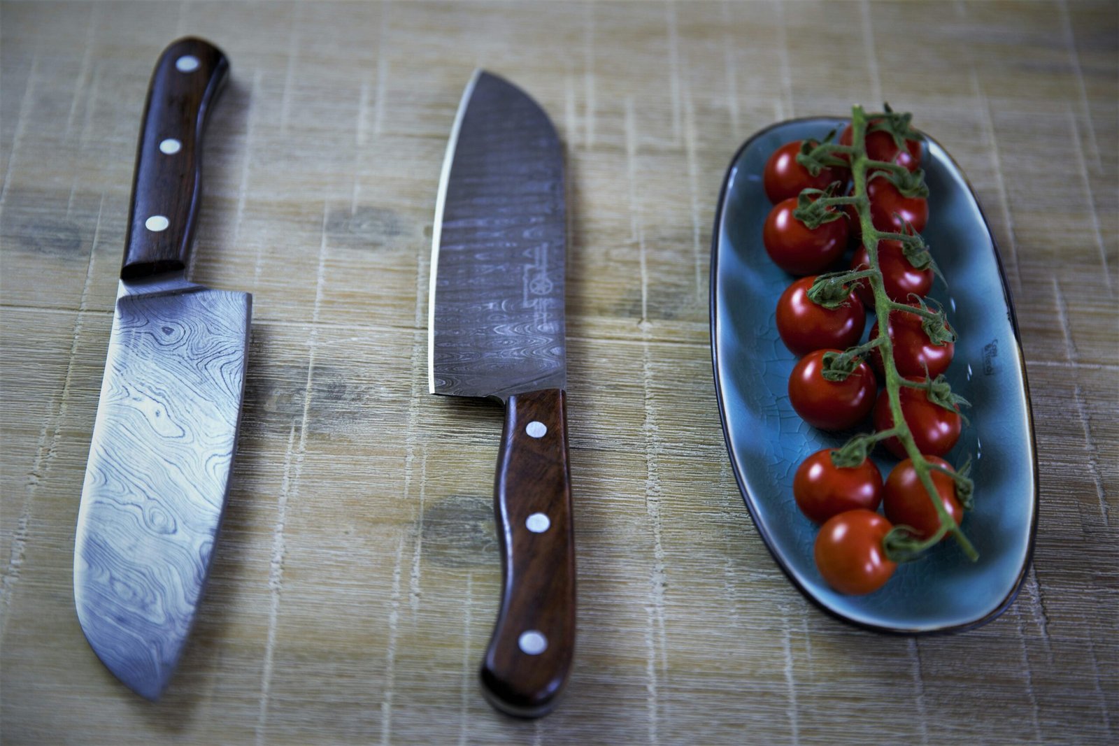 The image shows two knives with wooden handles placed on a wooden surface. To the right of the knives, a plate holds a vine of fresh cherry tomatoes. The knives have a distinct pattern on the blade, suggesting a high-quality finish.